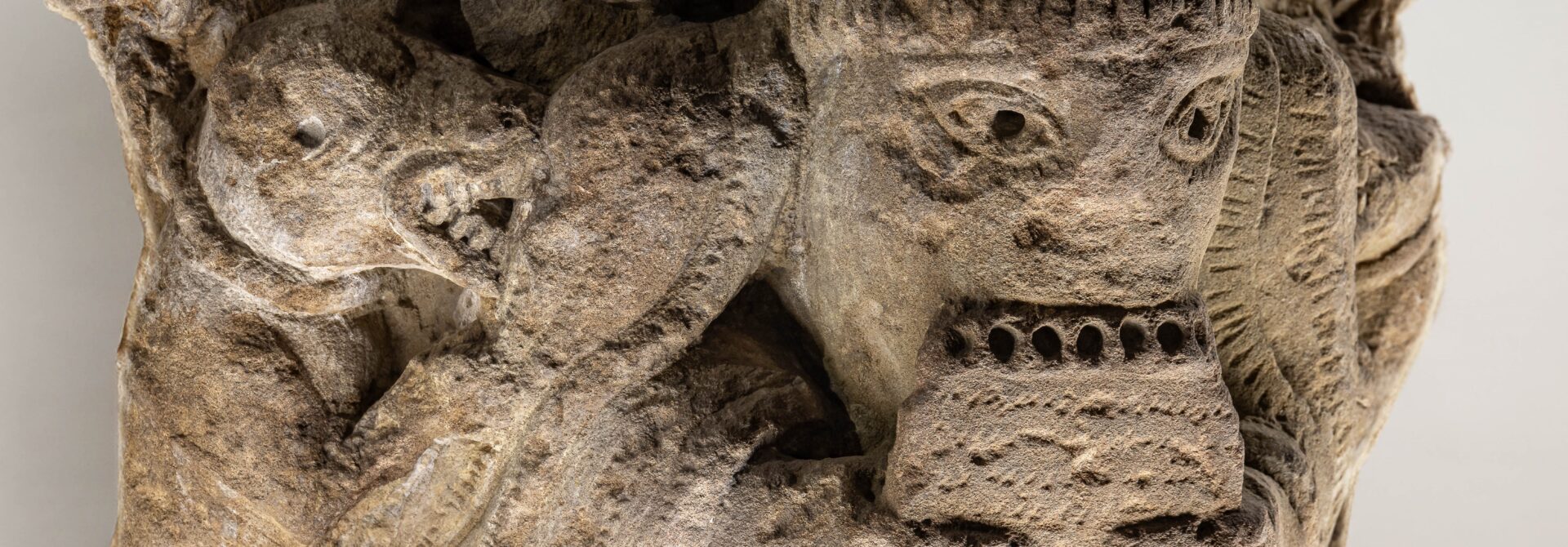 Gros plan sur un chapiteau figuratif en pierre présentant un personnage anthropomorphe stylisé.